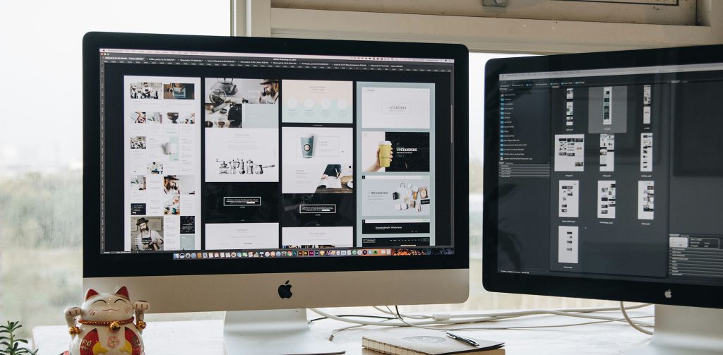 Ein Schreibtisch mit zwei Apple-iMac-Computern, auf deren Bildschirmen eine grafische Benutzeroberfläche und Design-Mockups zu sehen sind. Im Vordergrund stehen eine japanische Maneki-neko-Winkekatze, ein weißes Keyboard, eine Maus, ein Notizbuch, ein Smartphone und eine kleine Zimmerpflanze. Der Schreibtisch befindet sich vor einem großen Fenster mit Blick auf eine städtische Skyline.
