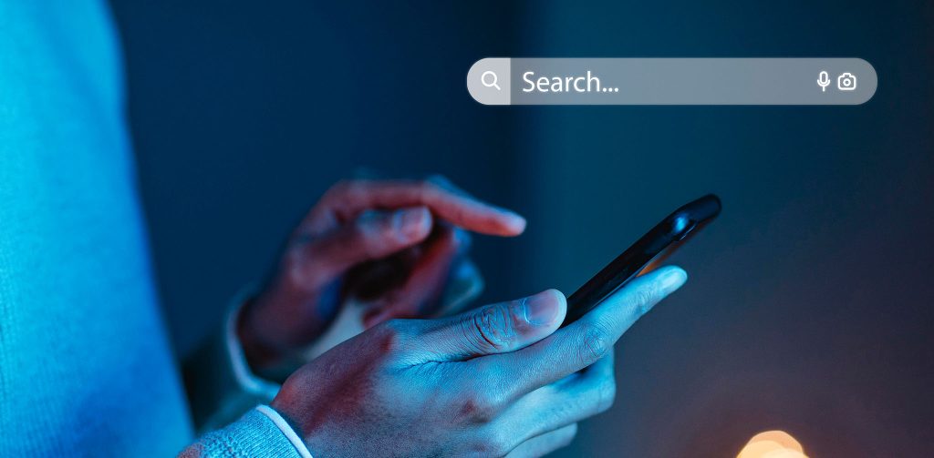 Detailaufnahme der Hände einer Person, die in einer dunklen, blau beleuchteten Umgebung ein schwarzes Smartphone mit dem Finger bedient. Am oberen Bildrand ist eine schwebende, hell leuchtende Suchleiste mit dem Text „Search...“ und einem Suchsymbol sowie einem Kamerasymbol eingeblendet. Die Person trägt ein helles, möglicherweise graues Oberteil mit Struktur. Im Vordergrund sind unscharfe, warme Lichter zu erkennen, die einen Kontrast zur kühlen, digitalen Atmosphäre bilden.