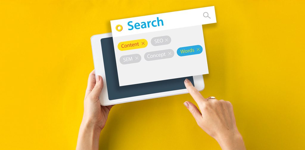 Eine Nahaufnahme von Händen, die einen weißen Tablet-Computer auf einem leuchtend gelben Hintergrund halten. Über dem Tablet schwebt ein Suchfeld mit dem Wort "Search" (Suchen) und einer Lupe. Darunter sind verschiedene Suchbegriffe als farbige Tags angeordnet, darunter "Content" (Inhalt) in Gelb, "Words" (Wörter) in Türkis sowie "SEO", "SEM" und "Concept" (Konzept) in Grau.